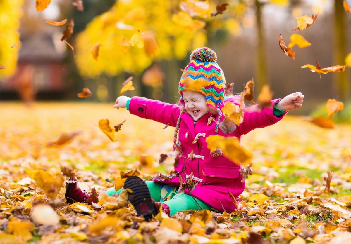 Happy little girl in pink warm coat and colorful knitted hat playing in beautiful autumn park on warm sunny fall day. Kids play with golden maple leaves. (Happy little girl in pink warm coat and colorful knitted hat playing in beautiful autumn park on
